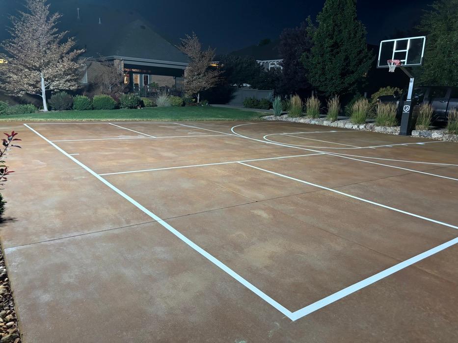 Multi-sport court at night on the Springville east bench, stained concrete striped in white for pickleball and basketball with a glass-backboard hoop.
