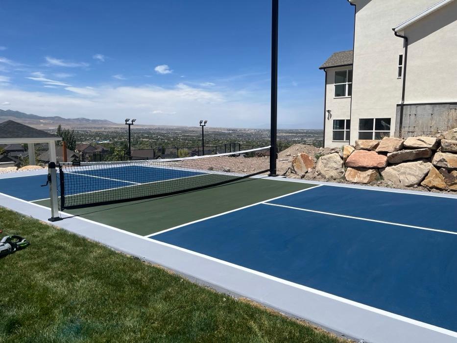Pickleball court on a bench lot above the valley in south Herriman, blue playing surface with a green non-volley zone and a boulder retaining wall along the high side.