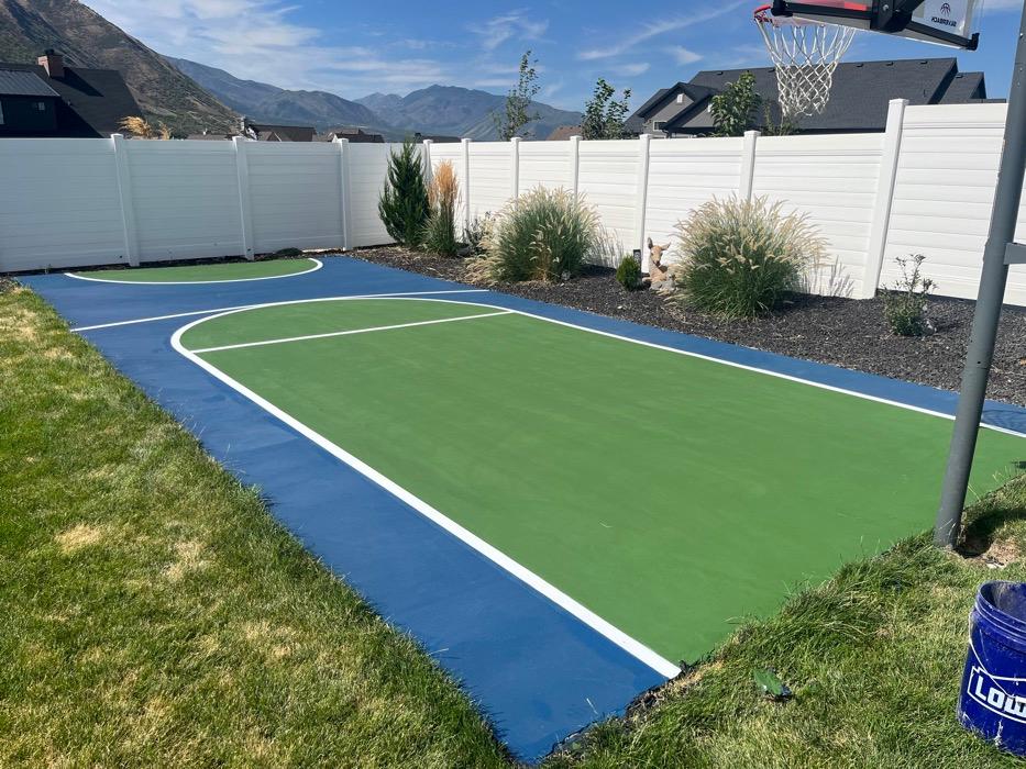 Backyard basketball half court in Spanish Fork, green key inside a navy surround with a pole-mounted hoop and white vinyl privacy fence.