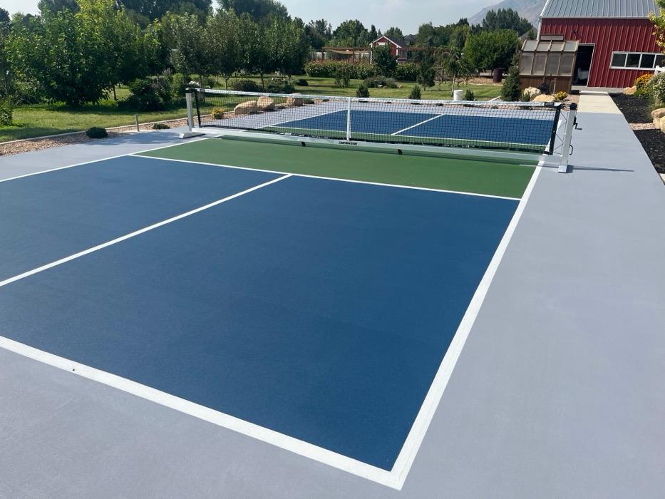 Pickleball court in Mapleton with a navy playing surface, grass green non-volley zone and light grey apron, orchard and a red barn beyond.