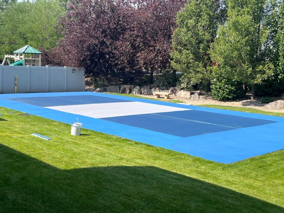 Two-tone blue pickleball court in Bountiful with a light grey non-volley zone, set into lawn with a boulder edge.