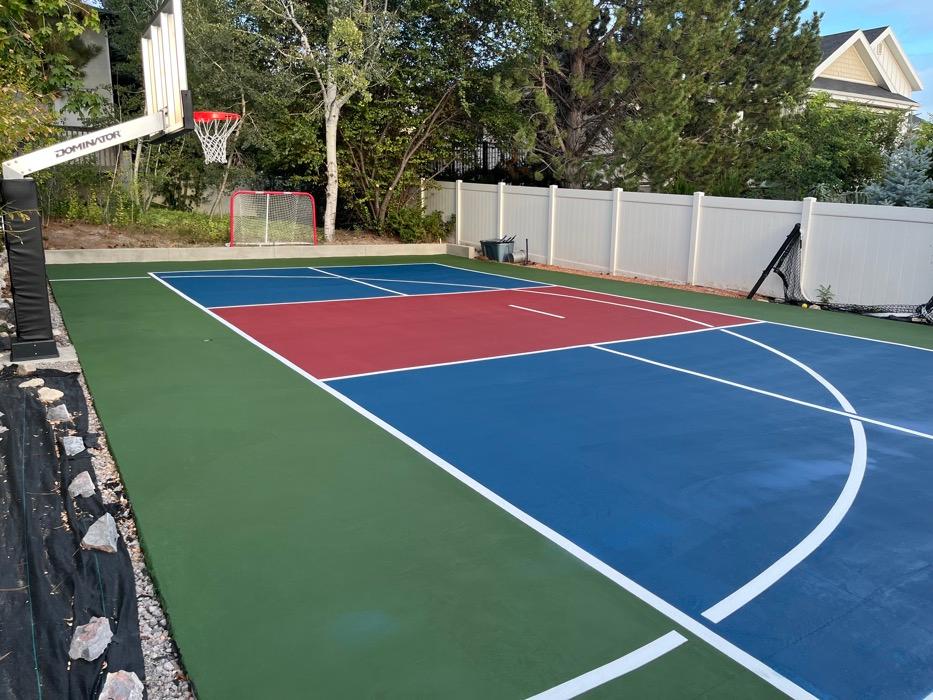 Multi-sport court in Pleasant Grove, red non-volley zone and navy zones inside a grass green apron, with a hoop and a concrete retaining wall at the far end.