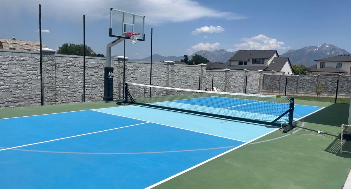 Multi-sport court in Cottonwood Heights built up to an existing stone wall, sky blue zones with a green apron and snow still on the peaks behind.