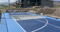 Multi-Sport Court Construction in Bear Lake, Utah looking toward the house built by Courts Utah your premiere multi-sport court contractor. This court has pickleball and basketball lines, a dominator basketball hoop, and black vinyl chainlink fencing.